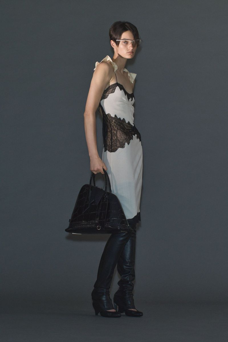 Model Tanya Churbanova wearing a white slip dress with black lace detailing, black knee-high boots, and holding a black handbag.