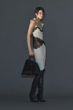 Model Tanya Churbanova wearing a white slip dress with black lace detailing, black knee-high boots, and holding a black handbag.