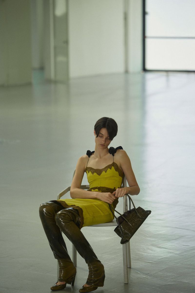 Model seated on a chair in a chartreuse slip dress and patent over the knee boots holding a handbag in an open studio space