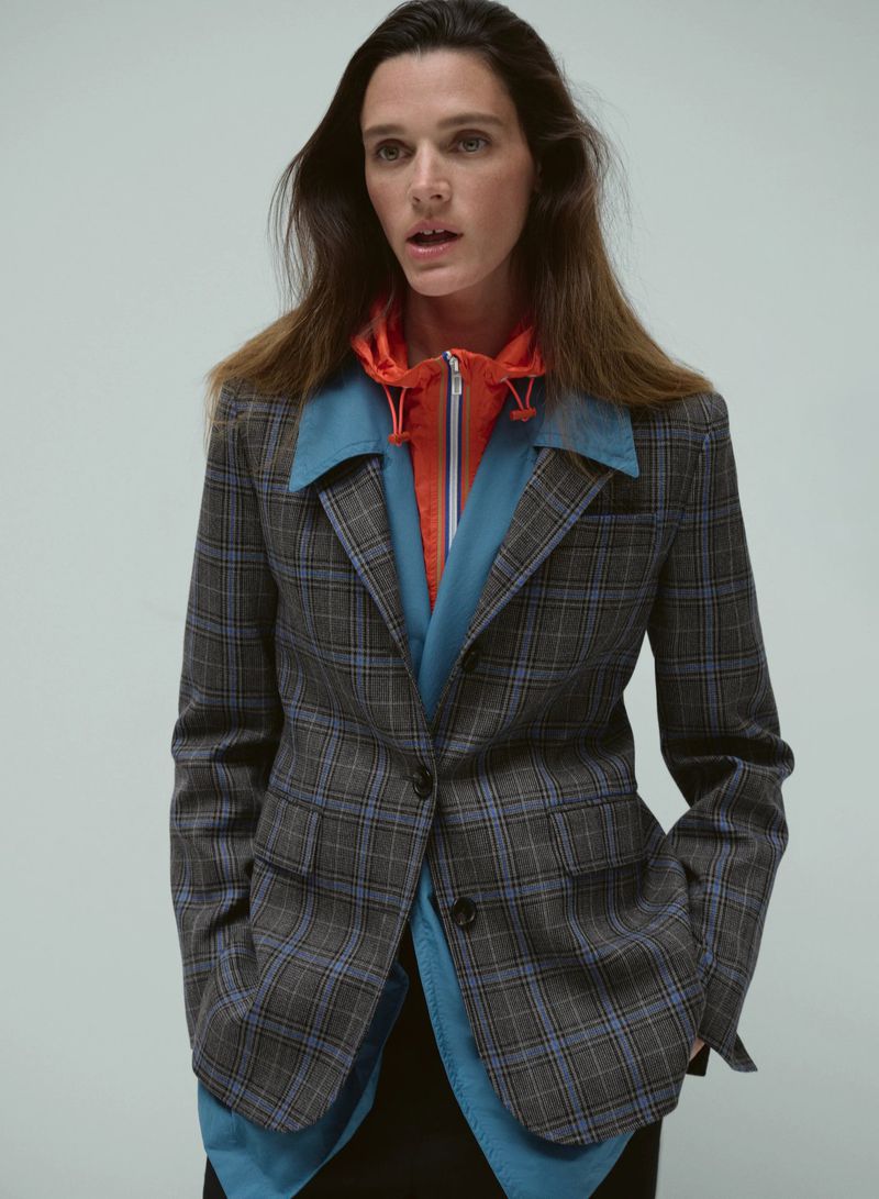 Model wearing a grey plaid blazer over a blue shirt and orange hoodie against a plain light grey backdrop