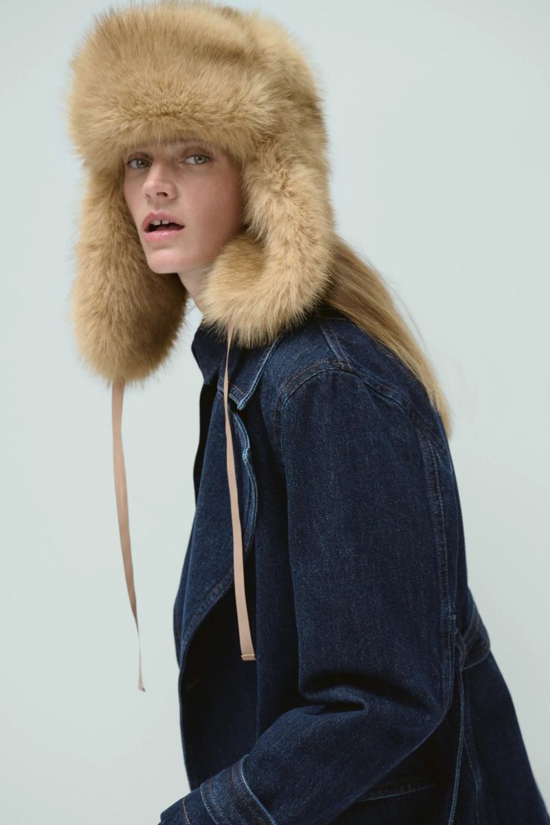 Leila Goldkuhl wearing a plush camel faux-fur trapper hat and dark blue denim jacket against a pale studio backdrop.
