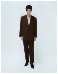 Model Billie Kennes posing in a brown oversized collarless pantsuit and black pointed-toe heels on a white seamless backdrop.