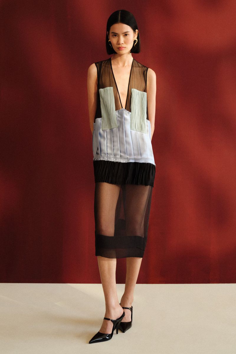 Model wearing a sheer sleeveless dress with pastel panels, black fringe, and pointed mules against a deep red backdrop.