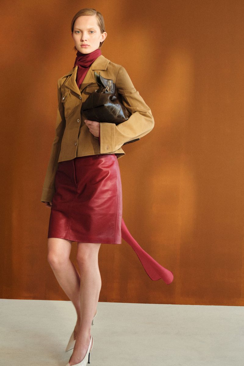 Model wearing a tan tailored jacket and burgundy leather skirt holding a dark brown bag against a warm backdrop.