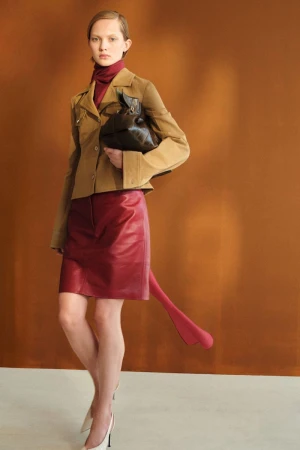 Model wearing a tan tailored jacket and burgundy leather skirt holding a dark brown bag against a warm backdrop.