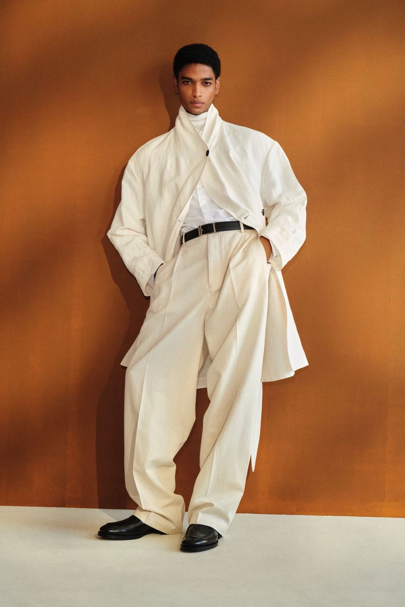 Male model wearing an oversized off-white wrap jacket and wide-leg trousers standing against a warm brown backdrop.