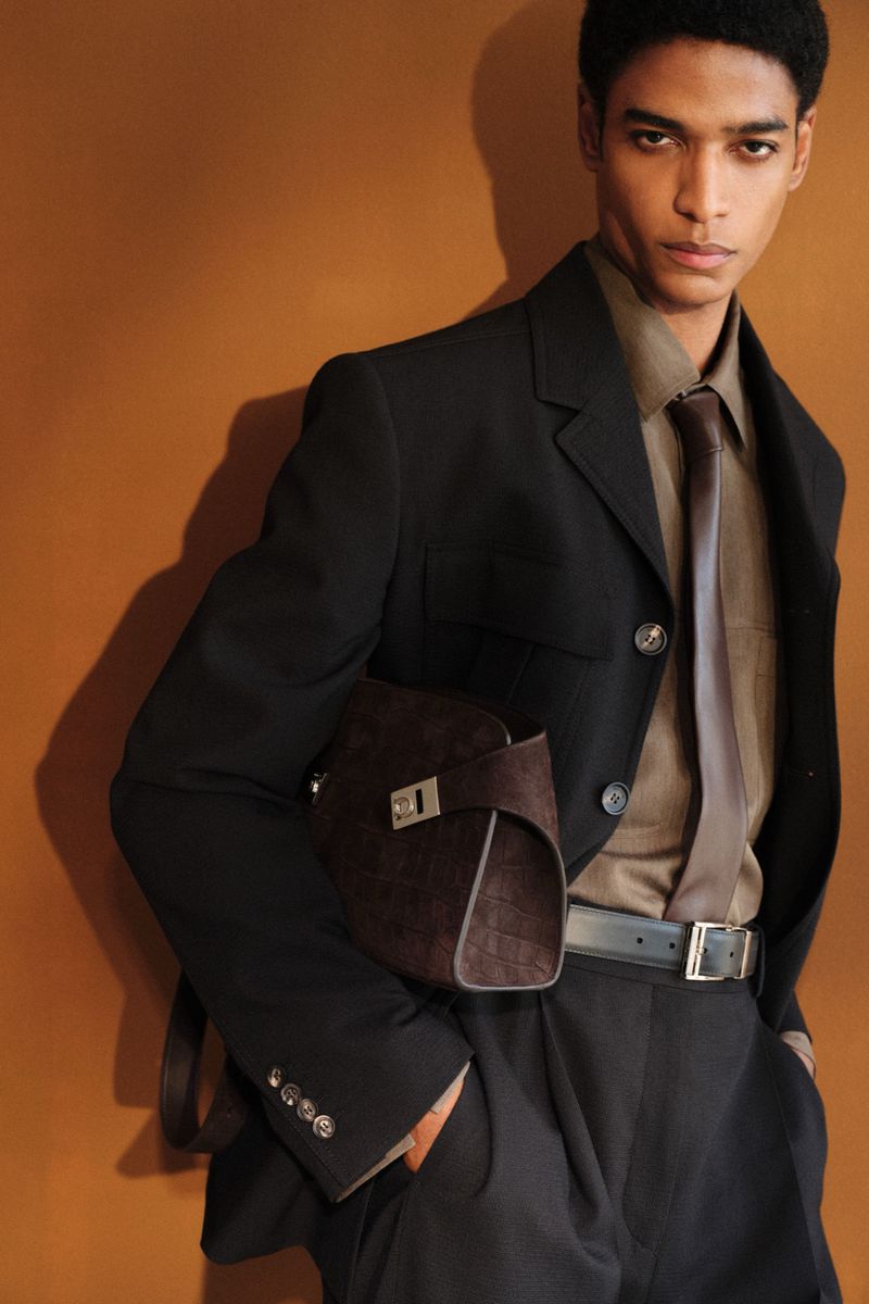 Male model in a black tailored suit and brown shirt holding a textured leather handbag against a warm brown studio background.