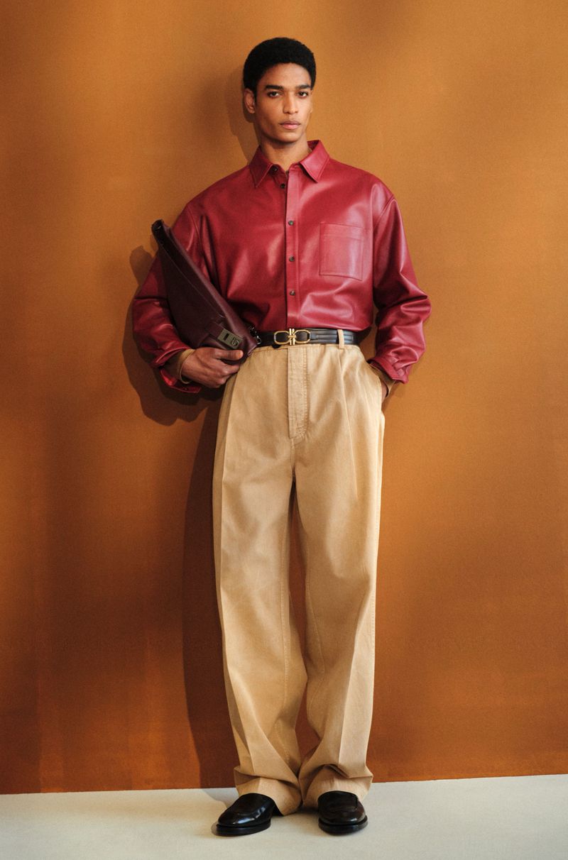 A male model in a red leather button-down shirt and beige wide-leg pleated trousers poses against a warm brown backdrop.