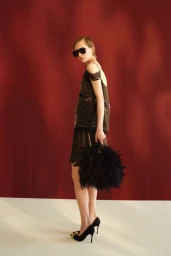 Model wearing dark sunglasses, an olive lace-trimmed mini set, and holding a black feather handbag against a crimson backdrop.