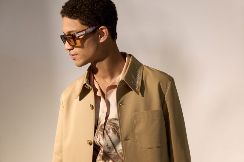 Male model wearing tinted sunglasses, an embroidered beige jacket, and a printed shirt against a neutral backdrop.