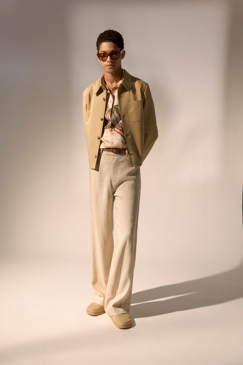 Model posing in a tan jacket, printed shirt, and cream wide-leg trousers with sunglasses against a neutral studio backdrop.
