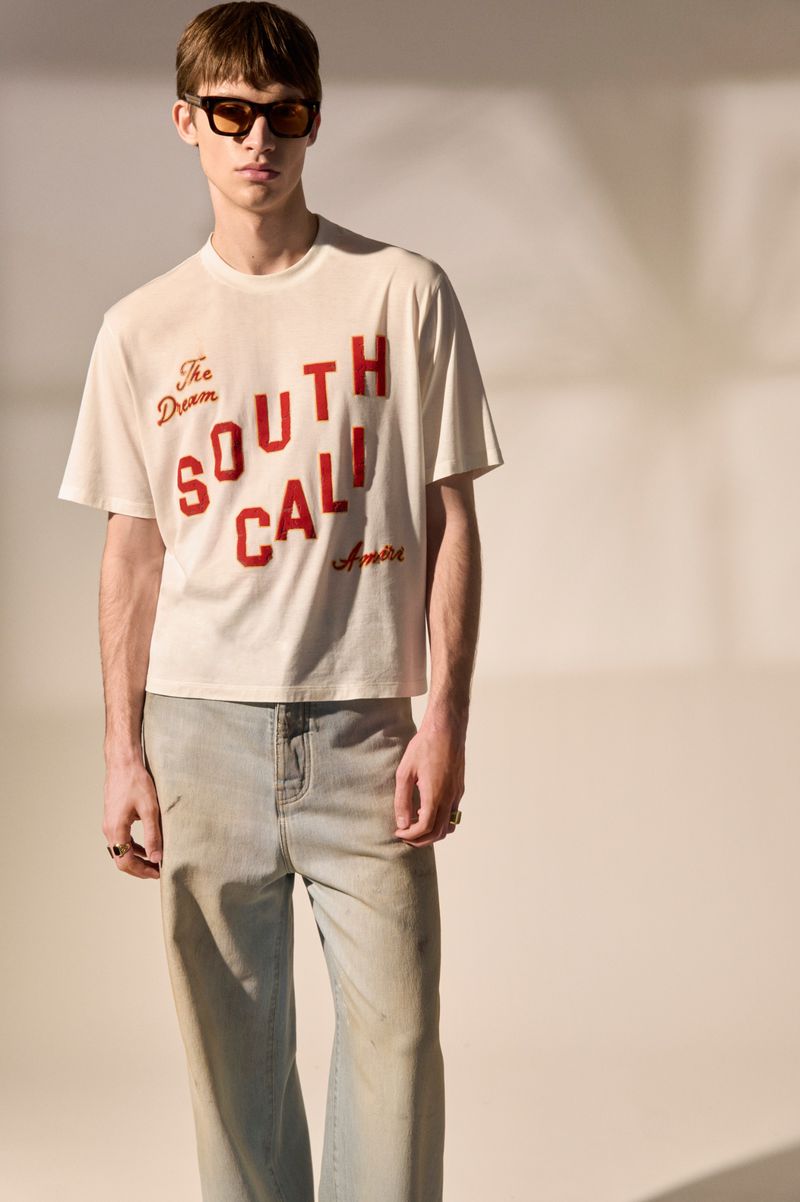 Male model in an off-white South Cali graphic t-shirt, light blue jeans, and dark sunglasses against a warm studio backdrop.