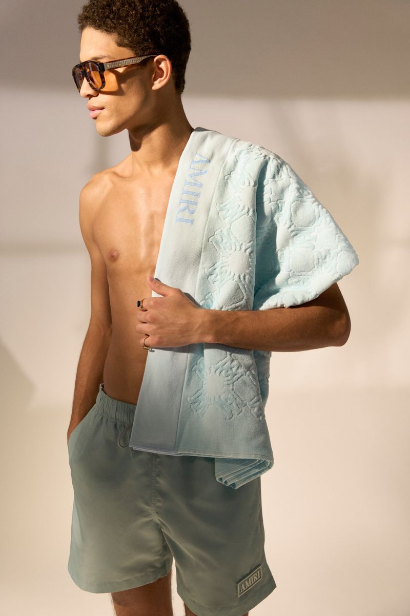 Shirtless male model wearing sunglasses, mint swim trunks, and holding a light blue Amiri towel over his shoulder in a studio.
