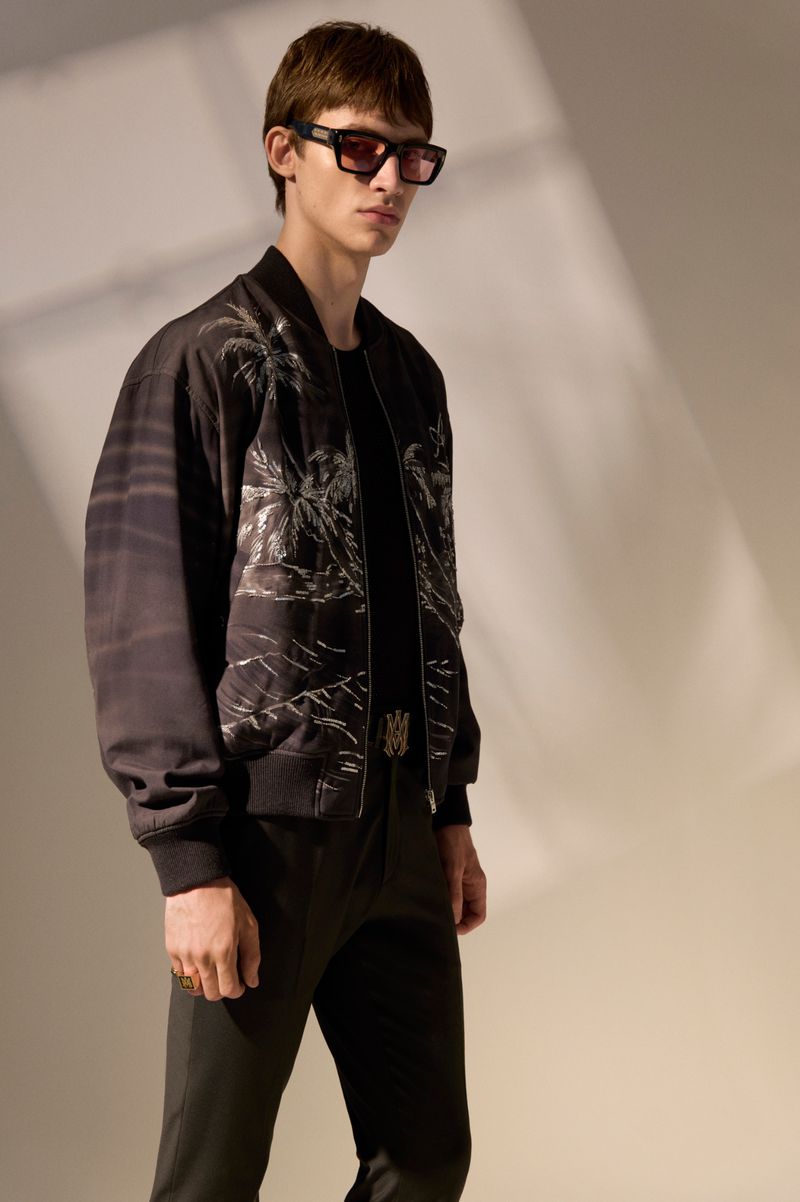 Model wearing dark sunglasses and a printed palm tree bomber jacket posing against a neutral studio wall with diagonal shadows.