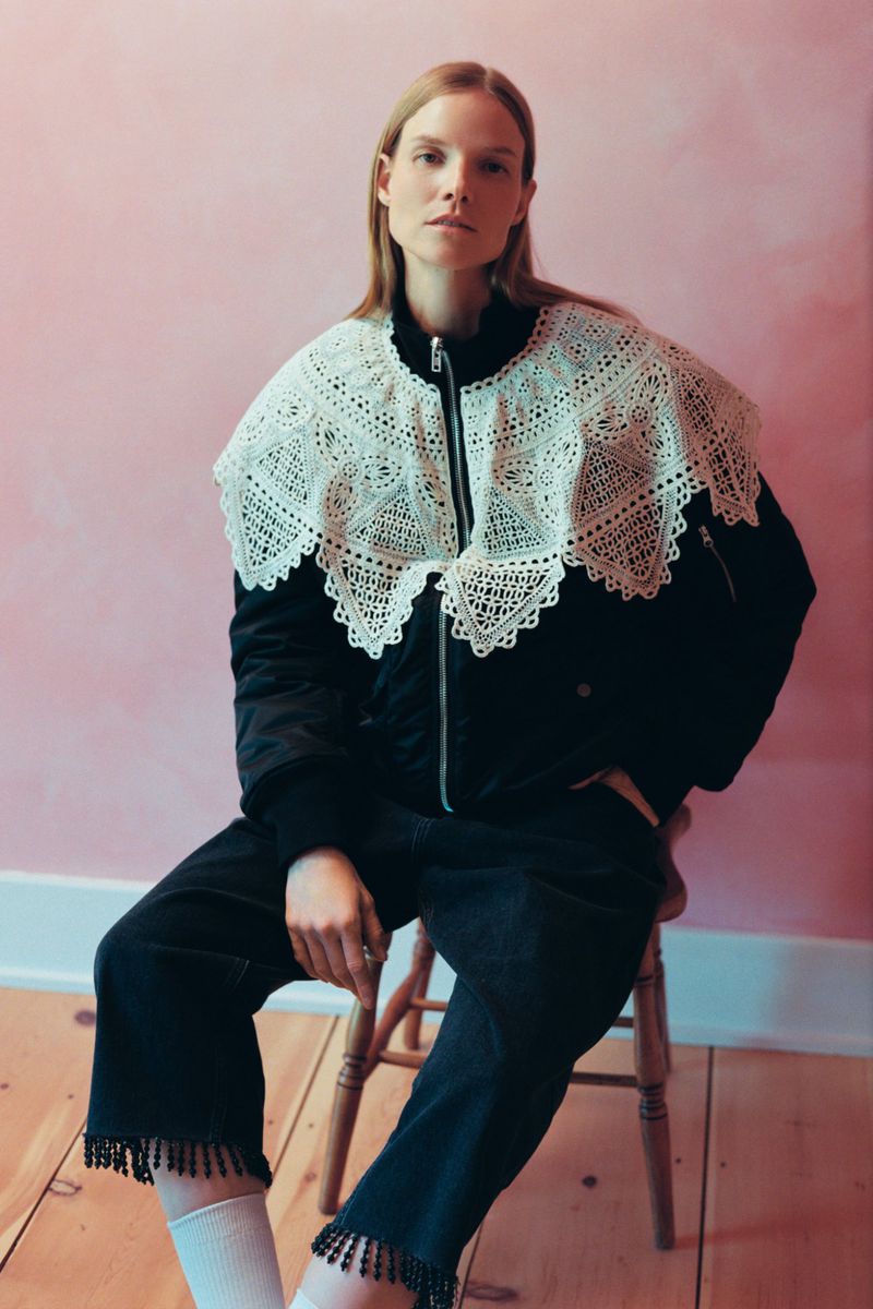 Model seated on a wooden chair wearing a black jacket with a large white lace collar and fringe-hem pants in front of a pink wall.