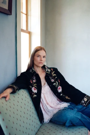 Suvi Riggs wearing a floral embroidered black jacket, blush top, and denim jeans while seated on a patterned sofa next to a window.