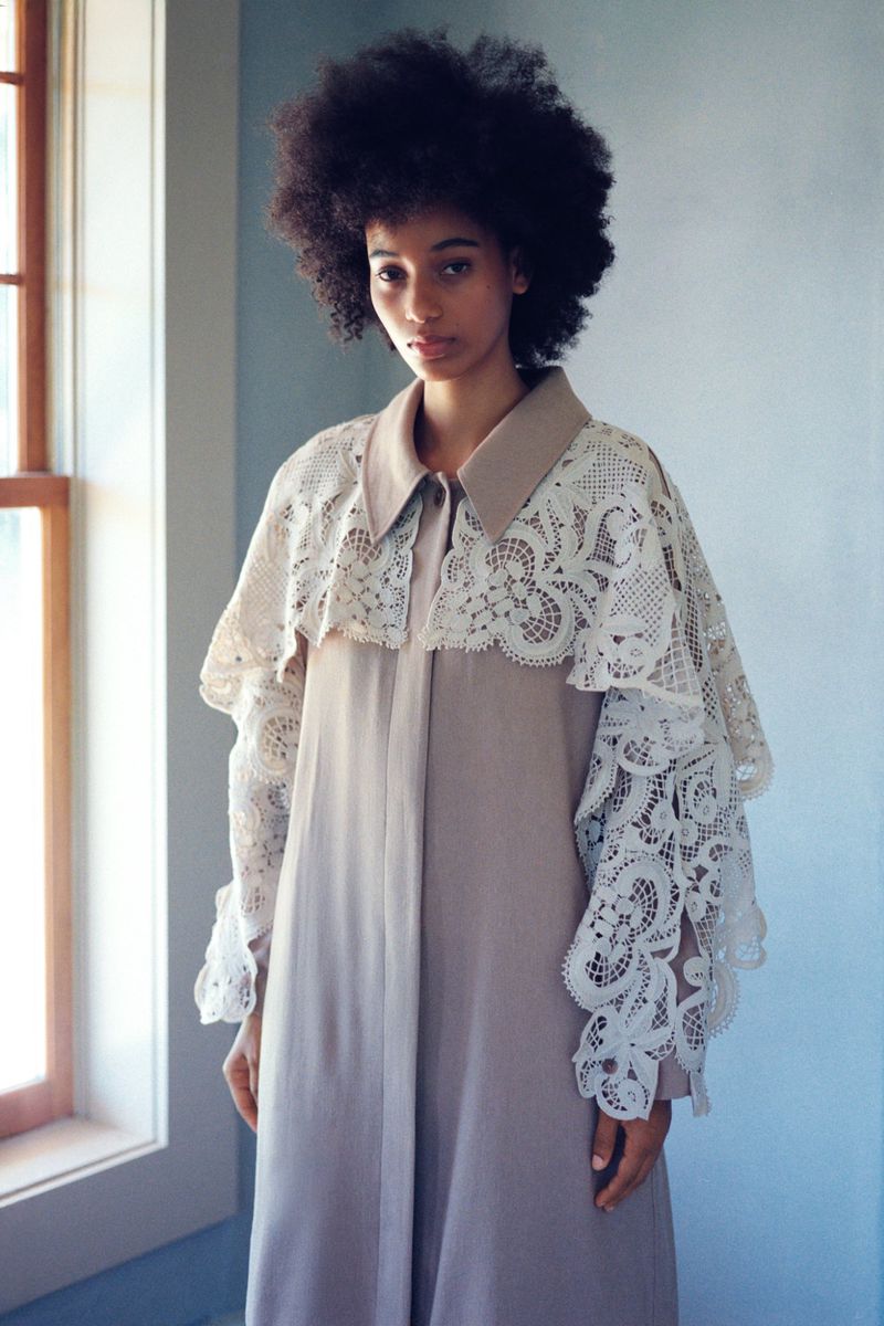 Fashion model wearing a taupe button-down coat with an ornate white lace cape collar standing beside a window.