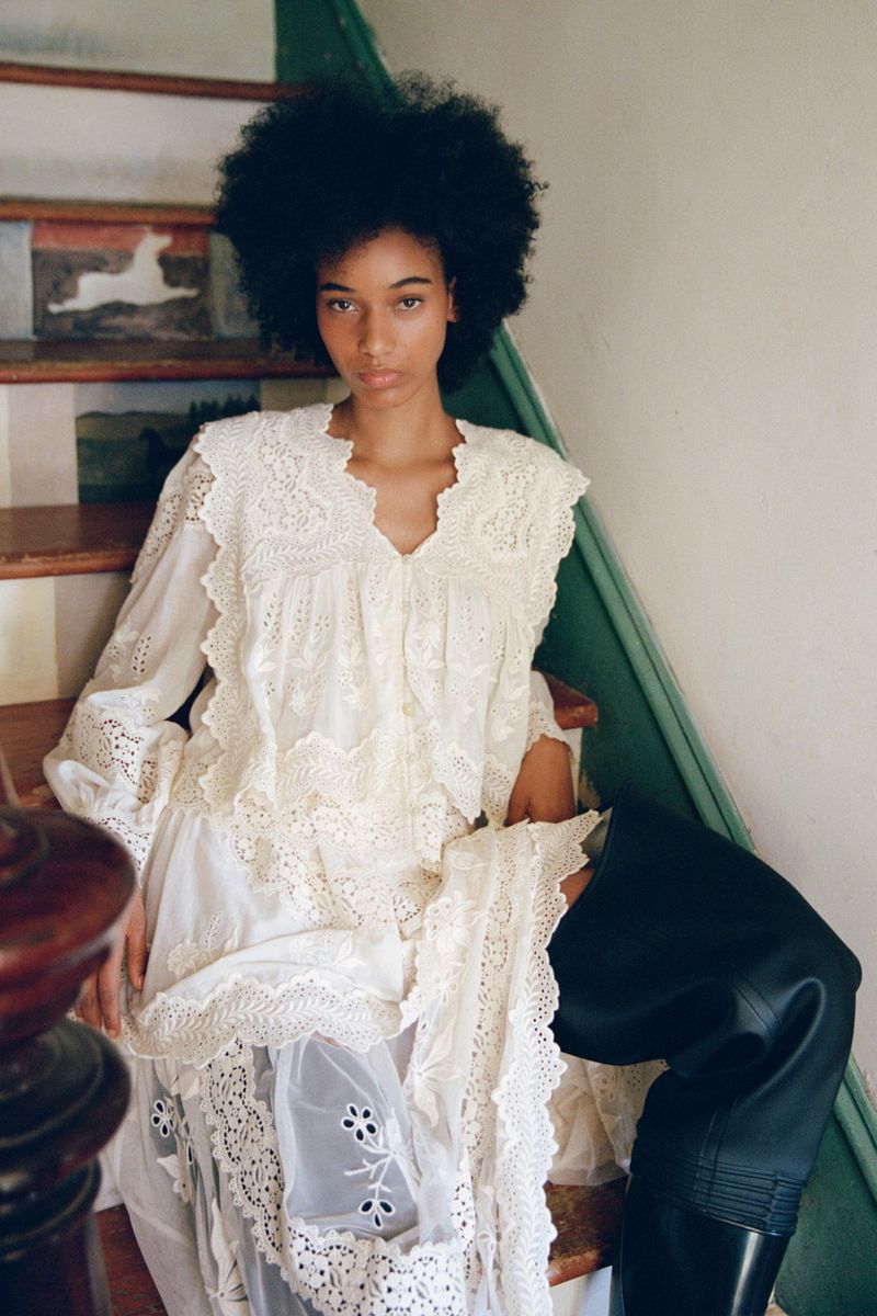 Model Manuela Sanchez seated on rustic wooden stairs wearing a white eyelet lace dress and tall black boots.