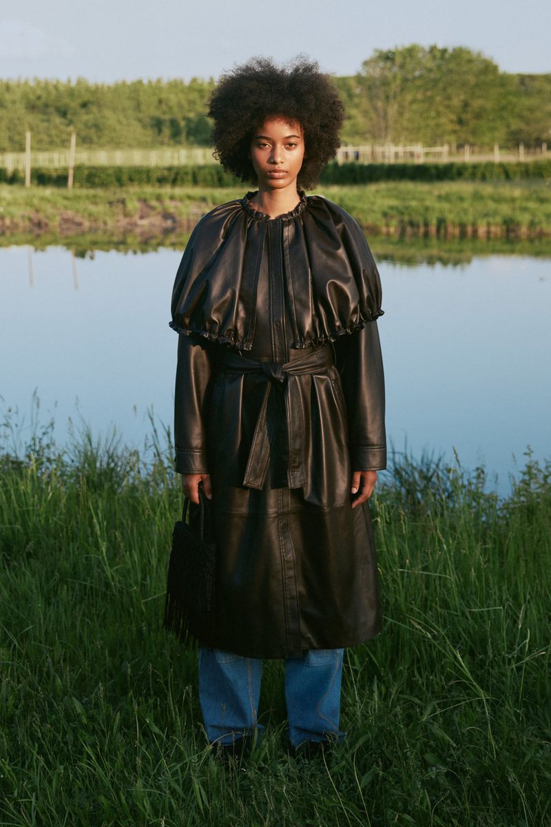Woman with afro hairstyle wearing a black leather ruffled cape coat and blue jeans standing in tall grass beside a calm lake.