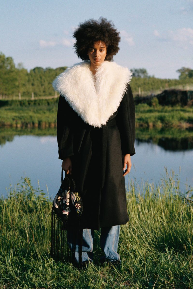 Model with afro hair in black coat with white fluffy collar and blue jeans standing outdoors near water