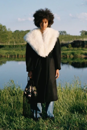 Model with afro hair in black coat with white fluffy collar and blue jeans standing outdoors near water