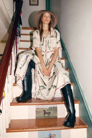 Model wearing printed dress, wide-brim hat, and tall boots, seated on indoor wooden stairs with painted risers.