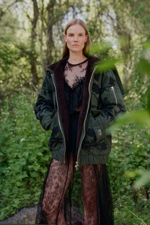 Model wearing an oversized dark green hooded bomber jacket over a sheer black lace dress in an outdoor woodland setting.