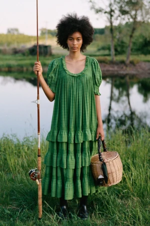 Model Manuela Sanchez wearing a tiered green puff-sleeve dress while holding a fishing rod and wicker basket by a pond.