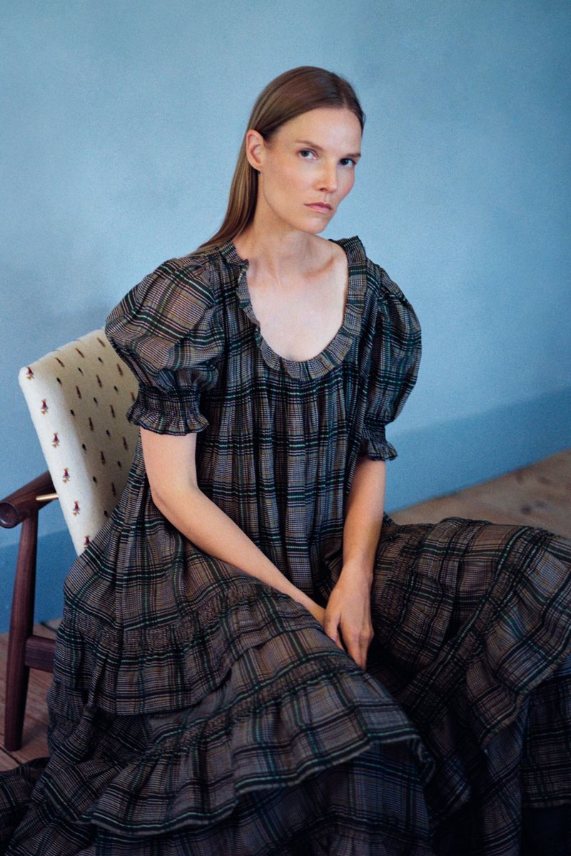 Model Suvi Riggs seated in a patterned chair wearing a voluminous plaid tiered maxi dress against a soft blue wall.