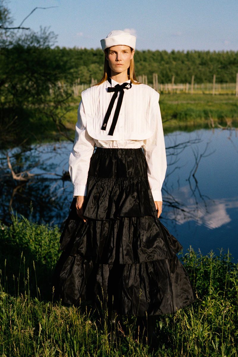 Suvi Riggs wearing a white sailor blouse, black tiered maxi skirt, and white cap standing near water for Sea RS27 Lookbook.