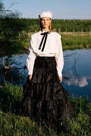Suvi Riggs wearing a white sailor blouse, black tiered maxi skirt, and white cap standing near water for Sea RS27 Lookbook.