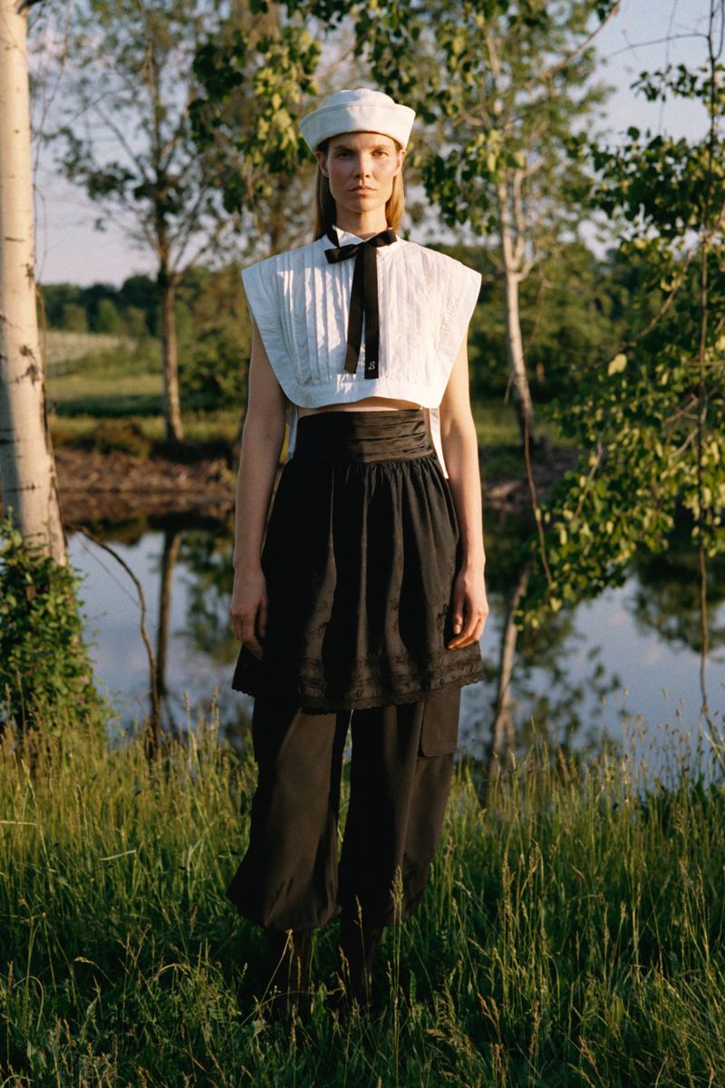 Model Suvi Riggs wearing a white cropped top, sailor hat, and layered black trousers in a grassy field by water for Sea.