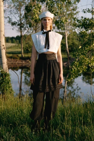 Model Suvi Riggs wearing a white cropped top, sailor hat, and layered black trousers in a grassy field by water for Sea.