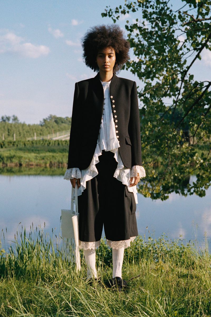Model with an afro wearing a tailored black blazer over a white ruffled top and black culottes standing on grass by a lake.