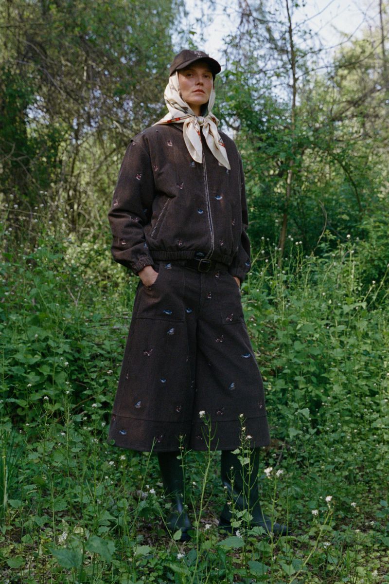 Model Suvi Riggs wearing a brown embroidered jacket, matching culottes, cap, and silk headscarf standing in a lush green woodland.