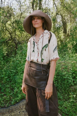 Suvi Riggs wearing a wide-brim brown hat, fish print shirt, and leather apron outdoors.