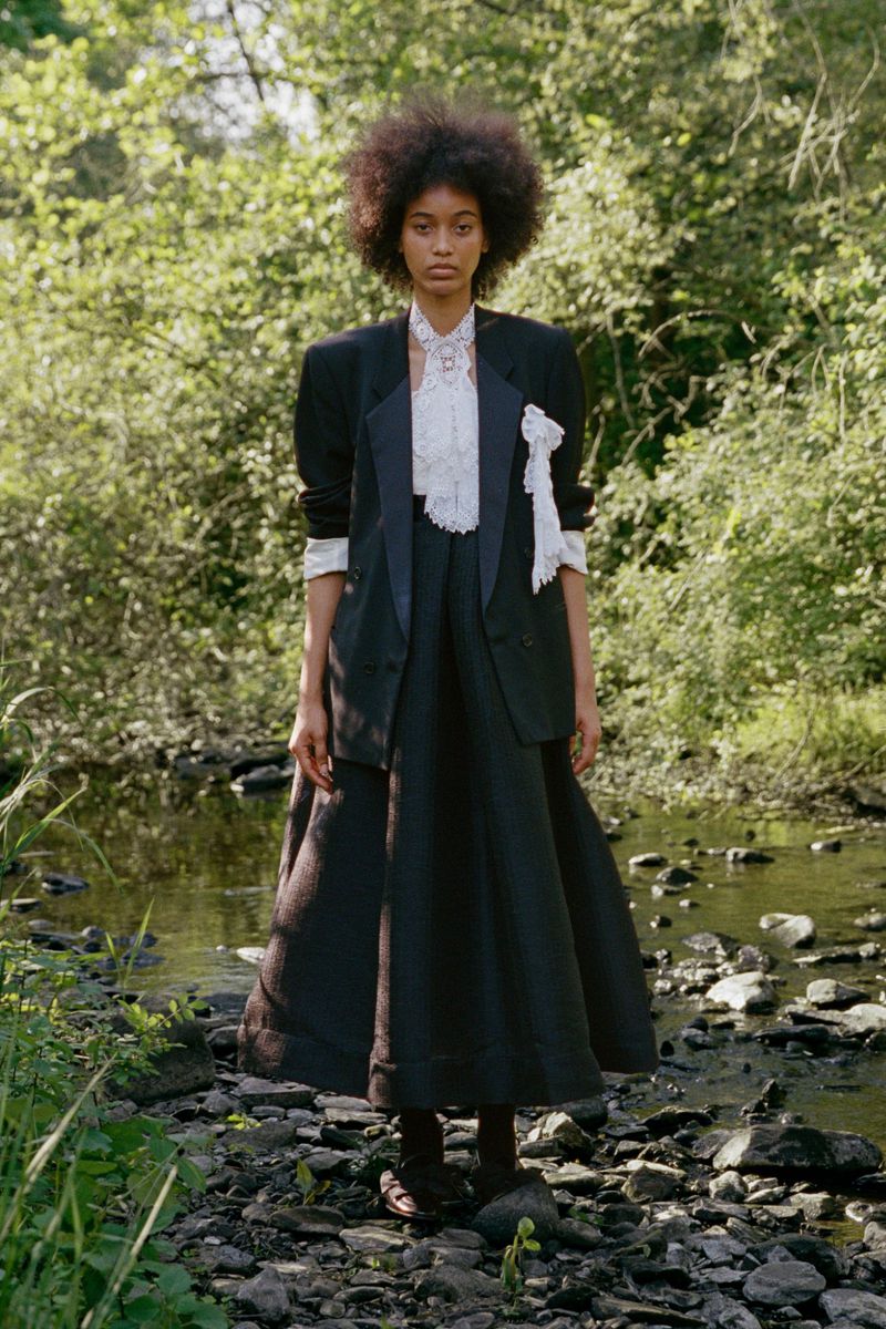 Model Manuela Sanchez standing in a rocky creek wearing an oversized black blazer, white lace ruffled blouse, and black maxi skirt.