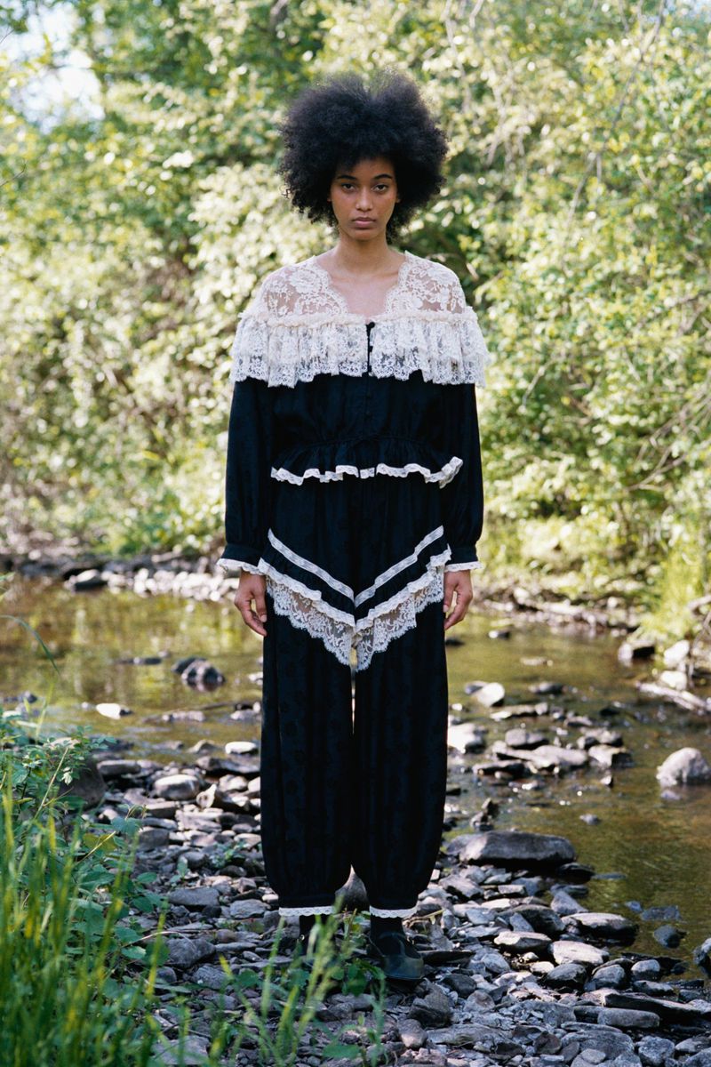 Model Manuela Sanchez in a black lace-trimmed two-piece ensemble standing on a rocky bank by a woodland stream.