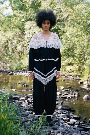 Model Manuela Sanchez in a black lace-trimmed two-piece ensemble standing on a rocky bank by a woodland stream.