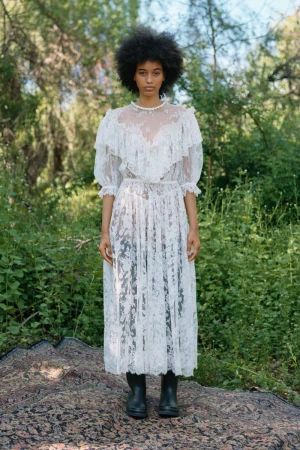 Model standing outdoors in a sheer white lace dress paired with black boots against a lush green wooded background.