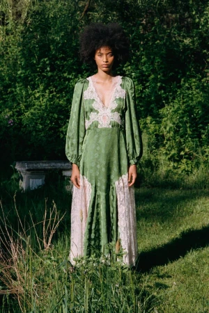 Model wearing a long green dress with white lace insets standing outdoors in tall grass before lush green foliage.