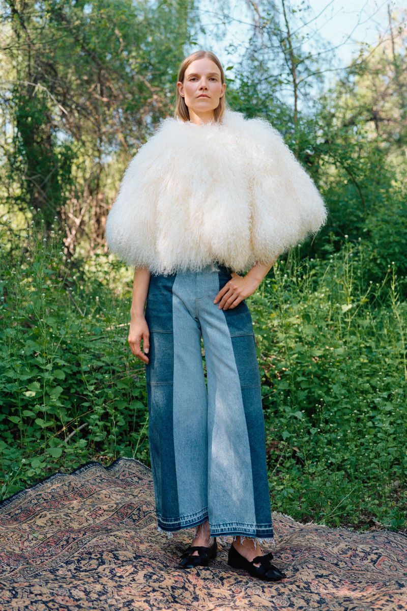 Model Suvi Riggs wearing a voluminous white fur cape and two-tone wide-leg denim jeans on a patterned rug in the woods.