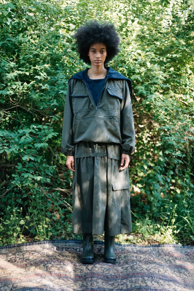 Model standing on an ornate rug outdoors in front of dense green foliage wearing an olive utility jacket and matching midi skirt.