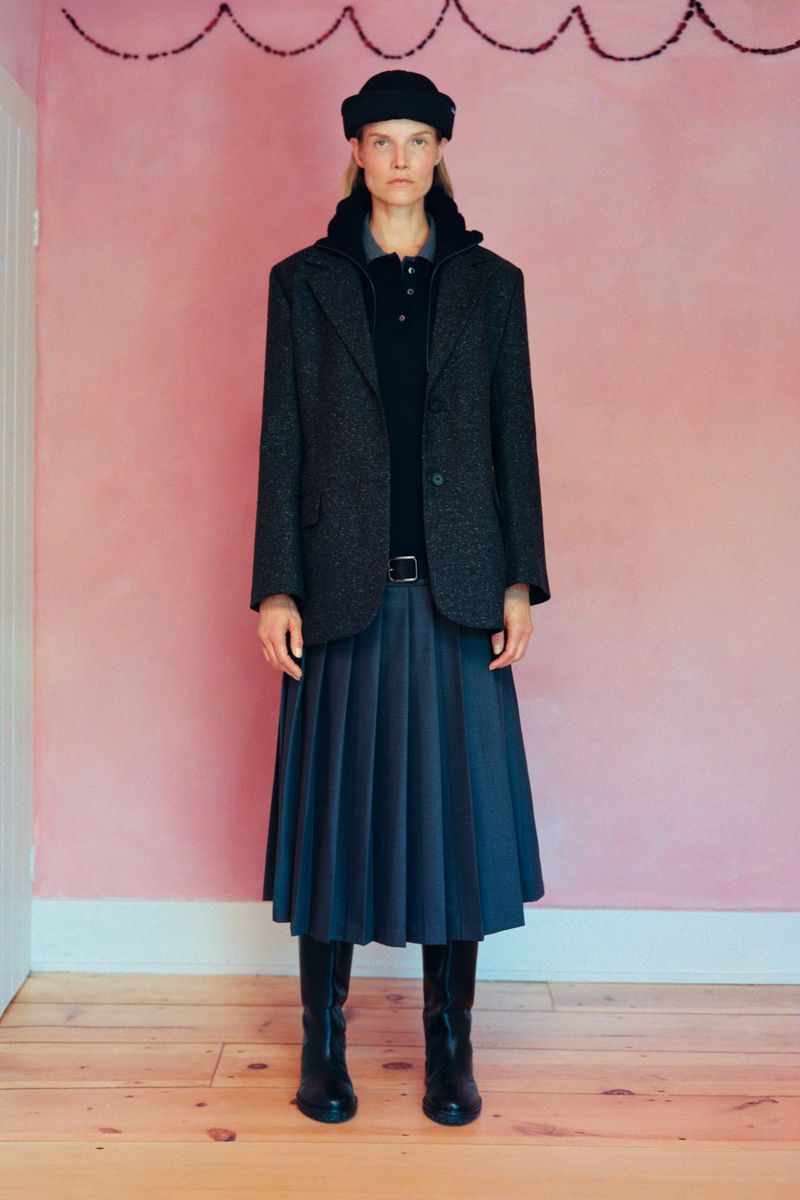Model Suvi Riggs wearing a dark wool blazer, pleated midi skirt, black beanie, and tall boots against a pink wall.