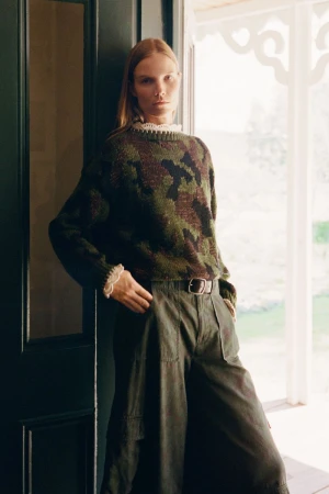 Model Suvi Riggs wearing a green camouflage knit sweater and wide-leg trousers in a doorway for Sea RS27 Lookbook.