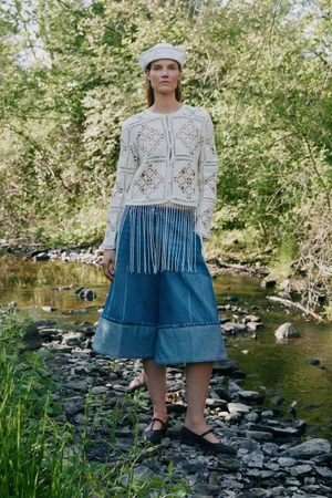 Model Suvi Riggs poses in a rocky stream wearing a white crochet cardigan with fringe, wide denim culottes, and a beret for Sea.
