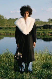 Model with afro hair in black coat with white fluffy collar and blue jeans standing outdoors near water