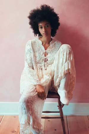 Model Manuela Sanchez with an afro wearing a cream lace tiered dress seated on a wooden chair against a pink wall.