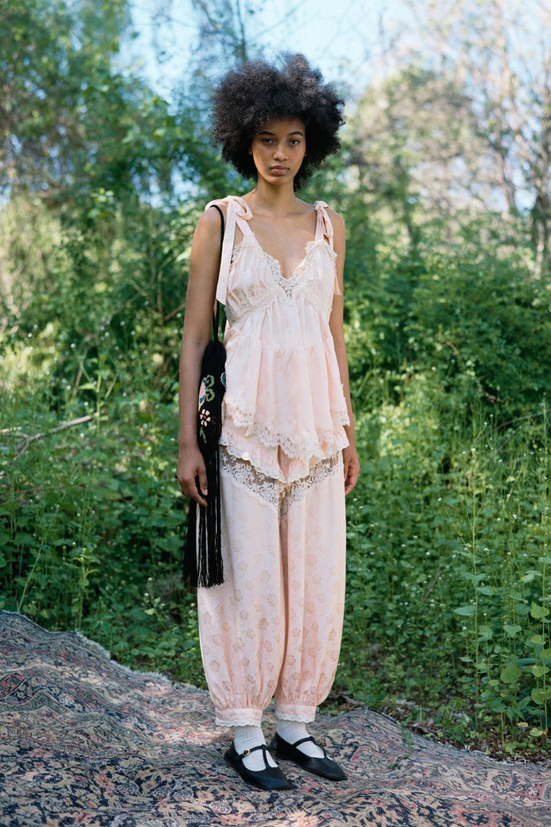 Model in a blush pink lace-trimmed top and matching bloomers standing on an ornate rug outdoors in front of lush green foliage.
