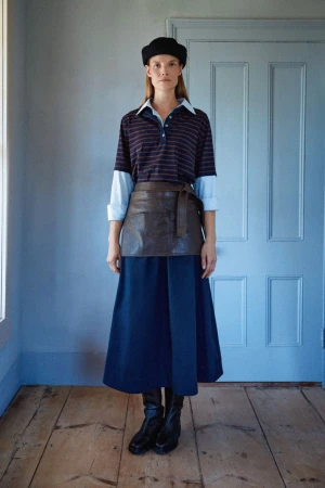 Model Suvi Riggs wearing a layered striped polo, blue midi skirt, brown wrap overskirt, and black beret standing before a paneled door.